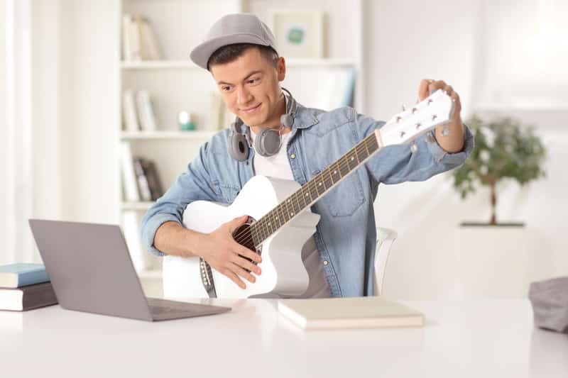 Guy sitting in front of a laptop computer at home and tuning a guitar