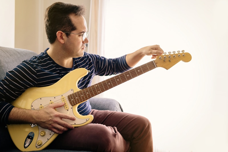 Boy tuning his electric guitar sitting on the sofa at home